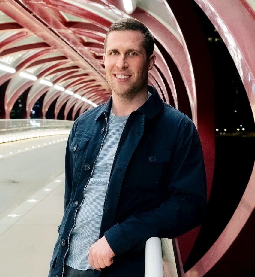 A man smiling and leaning on a railing, wearing a dark navy overshirt over a light gray t-shirt and dark pants, set against a modern architectural background with red and white curved structures.